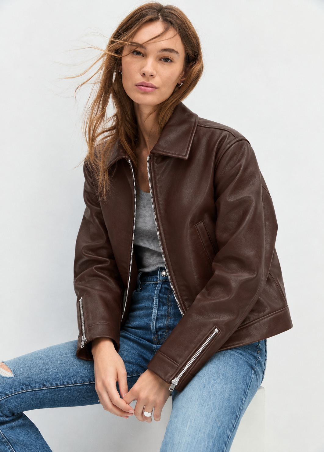 Woman wearing a brown cropped leather jacket open over a grey tank top with blue straight-leg jeans