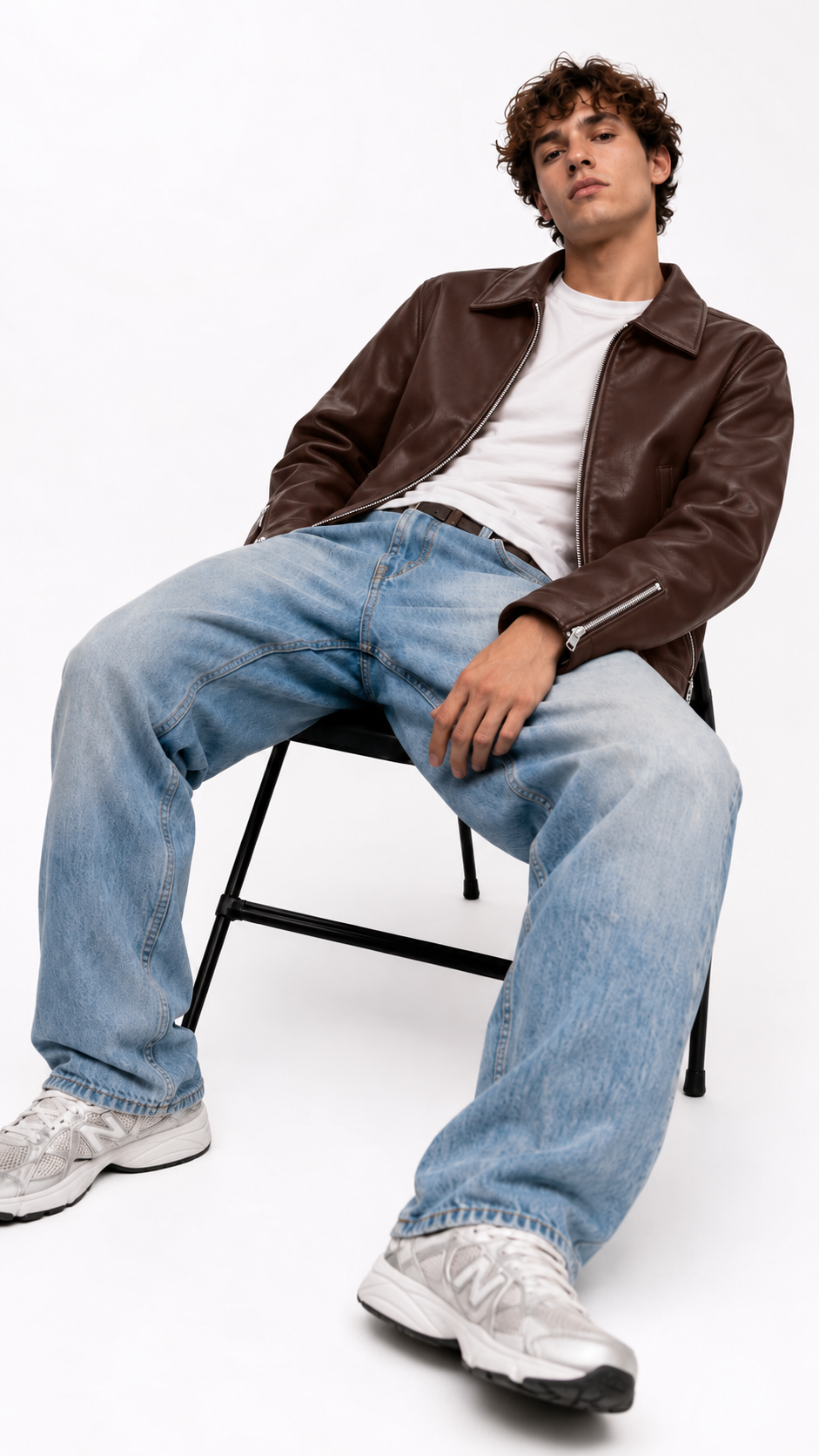 Man wearing a brown leather jacket open over a white tee with light-wash baggy jeans and grey sneakers